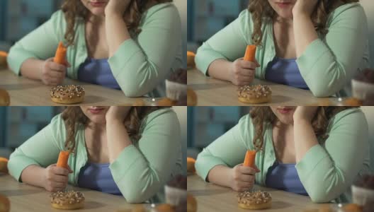 体重超标的女孩一边吃新鲜胡萝卜，一边悲伤地看着桌上的甜甜圈高清在线视频素材下载