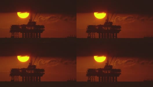 太阳从风暴云下与一个剪影的近海石油钻井平台在前景和石油(石油)油轮在背景在日落下面戏剧性的，暴风雨的天空高清在线视频素材下载