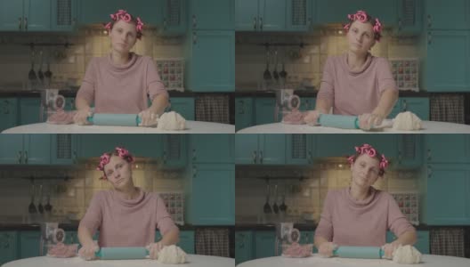 一名30多岁的年轻女子正用擀面杖擀面团，坐在厨房里看着摄像机。带卷发器在家独自做饭的家庭主妇。高清在线视频素材下载