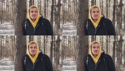 一个年轻人在森林里散步的肖像。一个穿着黄色连帽衫和黑色夹克的帅哥在早春倚在树上摆姿势高清在线视频素材下载