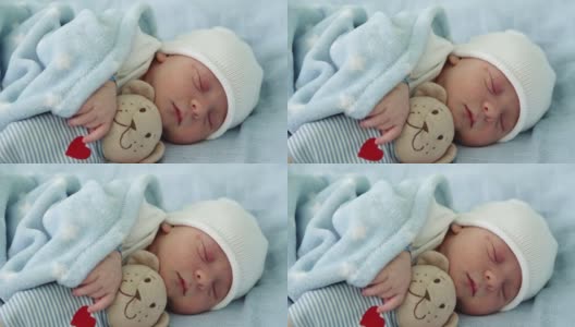 新生儿脸部特写，早期与泰迪熊睡觉的蓝色背景。在帽子上的生命开始分钟的孩子。婴儿，分娩，出生的最初时刻，开始的概念高清在线视频素材下载