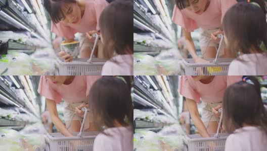 年轻的亚洲美丽的母亲拿着购物篮和她的孩子走在超市。她从货架上挑选绿色沙拉蔬菜，然后给她的小孩吃。健康的购物。高清在线视频素材下载