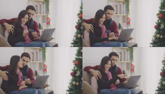 年轻的亚洲情侣情侣一起坐在沙发上。在以圣诞树为背景的房间里，用信用卡和笔记本电脑进行网上购物。4 k慢动作。高清在线视频素材下载