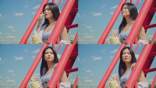 美丽的年轻亚洲女人吃爆米花，坐在公园的红楼梯在一个阳光灿烂的日子高清在线视频素材下载