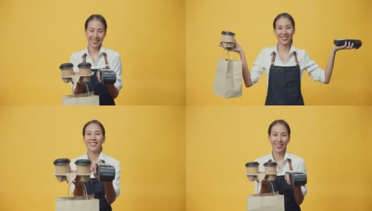 快乐微笑的亚洲女服务员穿着围裙友好地看着相机给顾客咖啡或茶和信用卡读卡机孤立在黄色背景。高清在线视频素材下载