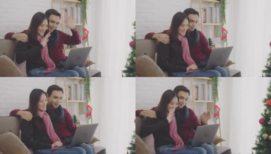 年轻的亚洲情侣情侣一起坐在沙发上。用笔记本电脑与家里装饰着圣诞树的房间里的家人和朋友进行视频通话。4 k慢动作。高清在线视频素材下载