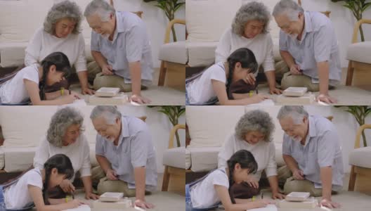 亚洲家族的祖父坐在客厅里笑着，用手摸着头表示爱。看孙女躺着写书，幸福老年退休高清在线视频素材下载