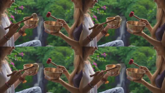 一名精通亚洲圣药的女子为一位年轻女性客户表演藏式碗治疗仪式的超慢镜头。用西藏唱碗冥想。他们在一个凉亭里冥想，背景是美丽的瀑布高清在线视频素材下载