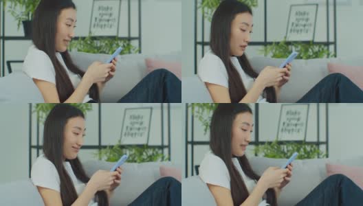快乐专注的混血儿年轻女子聊天用智能手机坐在舒适的沙发上。亚洲现代女性上网或在电话上打字。高清在线视频素材下载
