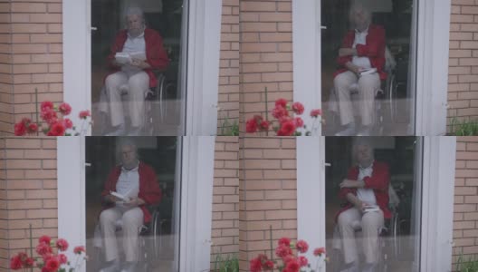 自信聪明英俊的老头坐在轮椅上看书合着书想着坐在家里的玻璃门后面。在室内用宽镜头拍摄的聪明体贴的白人退休老人。缓慢的运动。高清在线视频素材下载