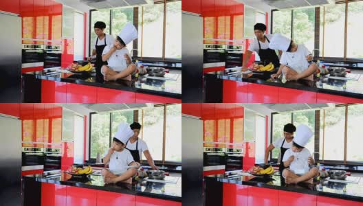 私人泰国厨师烹饪，他的小儿子在厨师帽旁边坐在桌子上的现代风格的家庭厨房。孩子在吃，爸爸在做饭高清在线视频素材下载