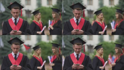 男人穿着学术服装和帽子在手机上浏览，女人在后台聊天高清在线视频素材下载