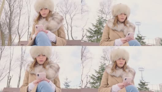 戴着皮帽的漂亮女人坐在公园长椅上，在冬天的城市公园里使用智能手机，在下雪的天气里，特写，慢镜头高清在线视频素材下载