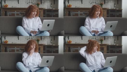 快乐的红发卷发女人用笔记本电脑聊天，坐在家里的沙发上通过在线视频聊天。高清在线视频素材下载