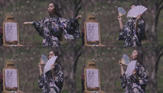 美丽的亚洲年轻女子穿着和服在春天的樱花花园与扇子跳舞微笑与画布画在背景模糊的画架。在阳光明媚的日子里，自信的日本女士的肖像。高清在线视频素材下载