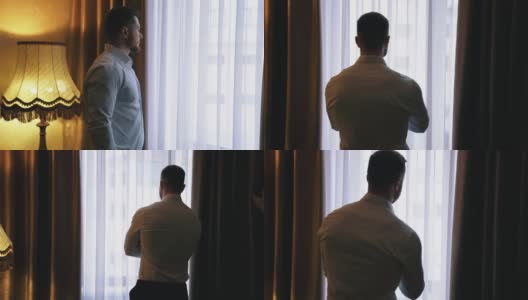 旅馆靠窗的男人。穿着白衬衫的年轻商人站在旅馆房间的窗户旁边，透过窗户往外看。后视图。高清在线视频素材下载