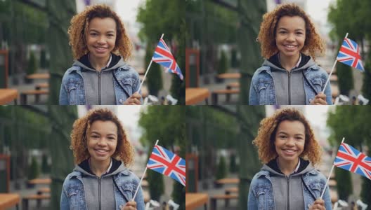 慢动作肖像年轻和快乐的非洲裔美国学生在英国举着英国国旗和微笑看着相机站在绿树的街道上。高清在线视频素材下载