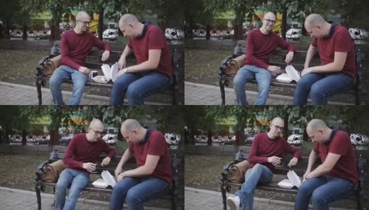 男人们坐在公园的长椅上吃着三明治和薯条，用杯子喝着咖啡聊天高清在线视频素材下载
