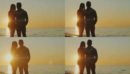 一对年轻的夫妇在海边拥抱着欣赏夕阳的剪影。后视图高清在线视频素材下载