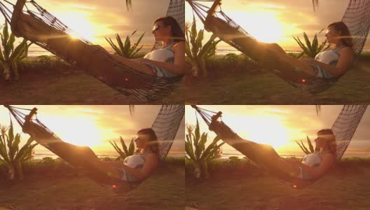 慢动作照明弹:黑发女子躺在摇摆的吊床上，在金色的日落海滩上高清在线视频素材下载