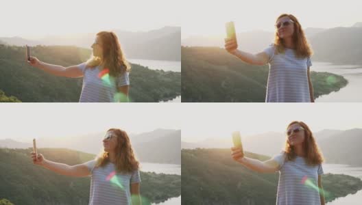 年轻的红发女游客戴着墨镜，站在高高的山顶上自拍，背景是一个巨大的山湖。放松。镜头光晕。旅行。生活方式高清在线视频素材下载