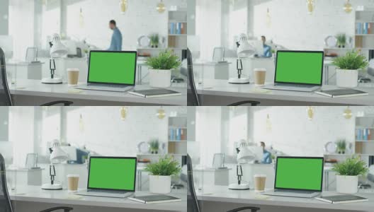 绿色屏幕的笔记本电脑站在桌子上，没有人在上面工作。背景里，一个男人走进来，坐在他的工作场所开始工作。高清在线视频素材下载