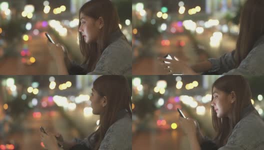 年轻美丽的亚洲女性微笑着使用智能手机上网社交媒体或网上购物，看着城市的夜光。漂亮女孩喜欢城市夜生活。高清在线视频素材下载