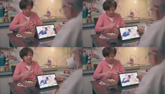 一位坐在轮椅上的亚洲老人在晚饭后的闲暇时间用数码平板电脑给家人打视频电话，而妻子则在厨房递给他苹果高清在线视频素材下载