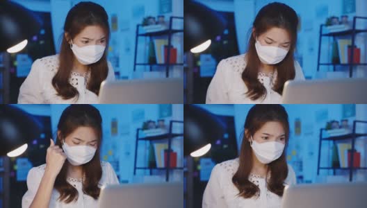 快乐亚洲女商人戴着医用口罩，在新常态下保持社交距离，以预防病毒，在办公室晚上使用笔记本电脑回到工作岗位。高清在线视频素材下载