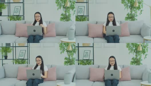 亚洲妇女戴着眼镜坐在沙发上戴着耳机使用笔记本电脑进行远距离通信。远程办公通过视频电话、辅导、在线会议等方式为客户提供支持。高清在线视频素材下载
