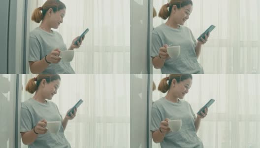 一名女子站在窗边，一边用智能手机一边在家喝热咖啡高清在线视频素材下载