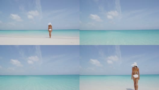海滩比基尼女人站在海边享受天堂海滩度假村。年轻的女士穿着白色比基尼和优雅的太阳帽在暑假。田园诗般的海滩景色，阳光灿烂的日子。高清在线视频素材下载