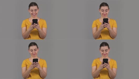 在灰色的背景下，身着黄色衬衫的年轻女子正开心地看着手机。社交媒体的概念高清在线视频素材下载