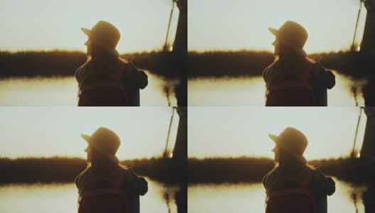 戴着帽子的快乐可爱女孩坐在夕阳下的河边码头上。金色的小时。享受美妙的旅行时刻。4K后视图特写高清在线视频素材下载