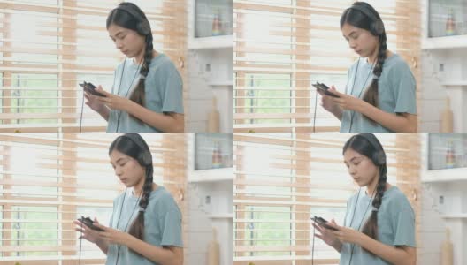 年轻美丽的亚洲女性戴着耳机和智能手机站在厨房，人和技术，Z一代的生活方式高清在线视频素材下载