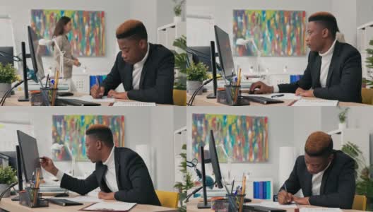 深色皮肤、身穿黑色西装的男子坐在电脑显示器前，描述打印在纸上的图形，与屏幕上的图形进行比较，指出，办公室同事在后台高清在线视频素材下载