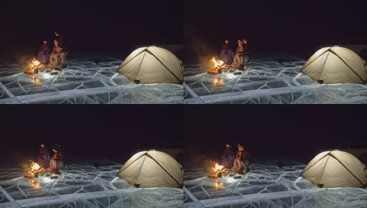 三个旅行者在夜间的冰面上着火。营地在冰上。帐篷紧挨着火。贝加尔湖。附近有一辆车。人们围着篝火取暖，穿着睡袋。高清在线视频素材下载