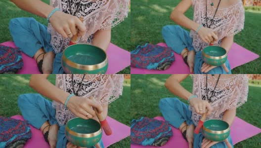 夏天，在公园里，一个女人坐在粉红色的瑜伽垫上玩歌唱碗。古董视。美丽的女孩拿着麻辣珠在绿草上沉思。慢动作高清在线视频素材下载