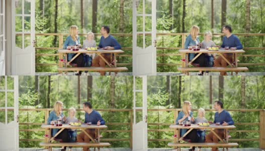 一个充满爱的家庭，母亲，父亲和小女儿在乡村别墅门廊的木桌上喝茶，谈笑风生高清在线视频素材下载