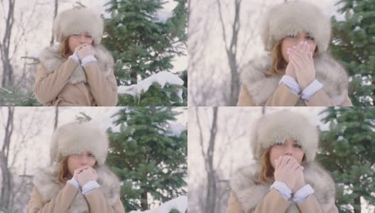 美丽的女人戴着皮帽看着镜头，试图温暖她的手在冬天公园在城市在下雪的天气下，慢镜头高清在线视频素材下载