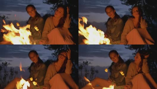 两个年轻的白人女孩晚上坐在篝火旁，准备一个棉花糖，看着篝火，思考着高清在线视频素材下载