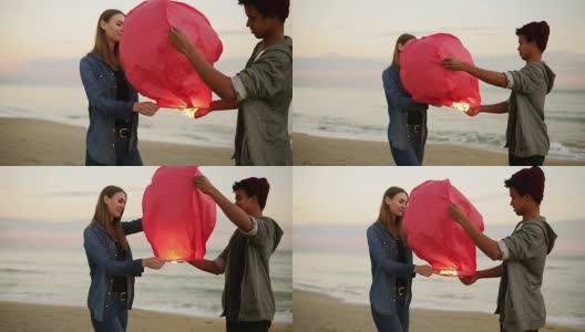 年轻的多民族夫妇举着红纸灯笼，准备出发。海滩上的浪漫约会。迷人的女人和她的非洲男朋友一起举着灯笼与火之前让它飞高清在线视频素材下载