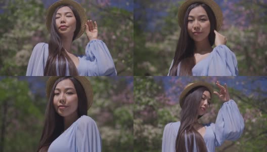 一个自信的亚洲女子，穿着蓝色上衣，手拿草帽，站在盛开的樱花公园，在阳光明媚的春日里看着镜头。优雅美丽的日本女士在花园里摆姿势的肖像。高清在线视频素材下载