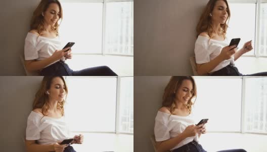 高兴的黑发女人戴着耳机坐在靠窗边听音乐和使用智能手机高清在线视频素材下载