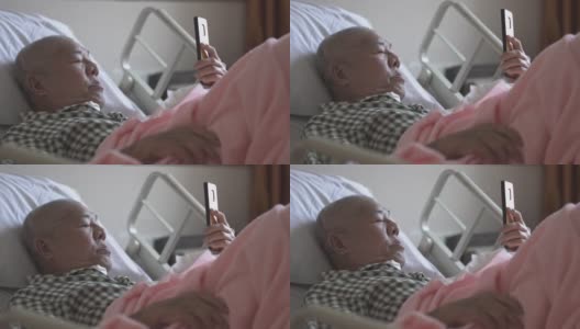 一位亚裔华裔老人躺在医院病房的病床上，用智能手机与家人视频通话高清在线视频素材下载