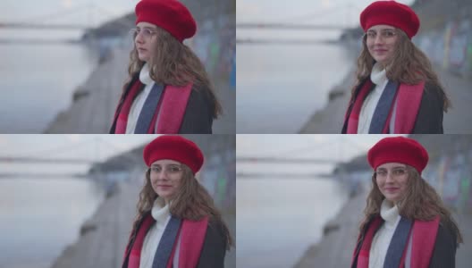 年轻迷人的女子在红色贝雷帽和眼镜钦佩美丽的自然站在城市的堤岸转向看着镜头微笑。周末户外高加索千禧一代的肖像。高清在线视频素材下载