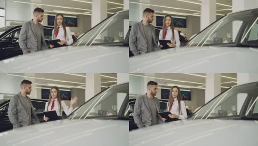 有魅力的女车商拿着文件站在漂亮的车旁边，正在和有兴趣的客户谈论新车型。高清在线视频素材下载