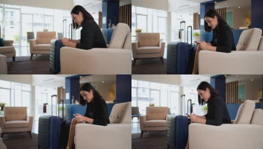 在一家豪华度假酒店的大厅里，亚洲女商人穿着正装，带着滚轮行李箱，一边在智能手机上输入信息，一边等着一位同事高清在线视频素材下载