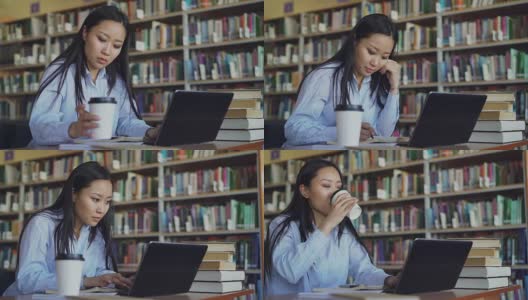 年轻的亚洲学生女孩坐在大学图书馆的一堆书的桌子上，在笔记本电脑上工作，喝咖啡微笑着看着别处，准备考试高清在线视频素材下载