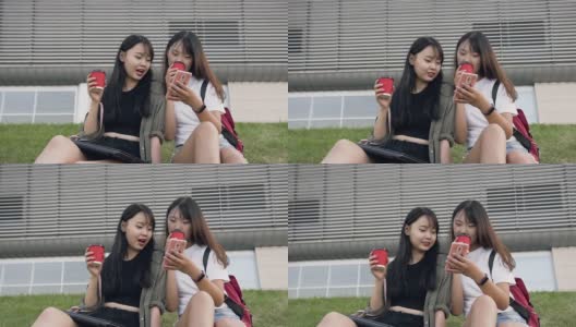 迷人的肖像两个愉快的亚洲褐发女人坐在草地上，使用他们的手机，喝咖啡在现代建筑的背景高清在线视频素材下载